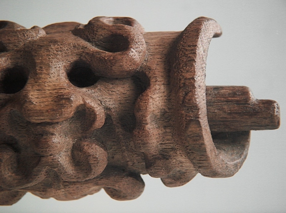 Rullverksornament.
Skulpterat litet, spänneliknande ornament, dekorerat i broskverksstil.
Det långsmala ornamentet är format som ett stycke kraftigt upprullad bit näver eller läder, med konkav bak- eller undersida. På baksidan är ornamentet profilerat för en list. Broskdekoren på ovansidan är formad som en maskaron.
Ornamentet är mycket välbevarat.

Text in English: A small, buckle-like ornament, decorated in a style similar to earshell work.
The long narrow ornament is formed as a piece of tightly rolled bark or leather, with a concave back or underside so as to match a batten or the like. The reverse side is profiled to fit a moulding. The earshell work on the upper side is shaped as a mask.
The ornament is well preserved.