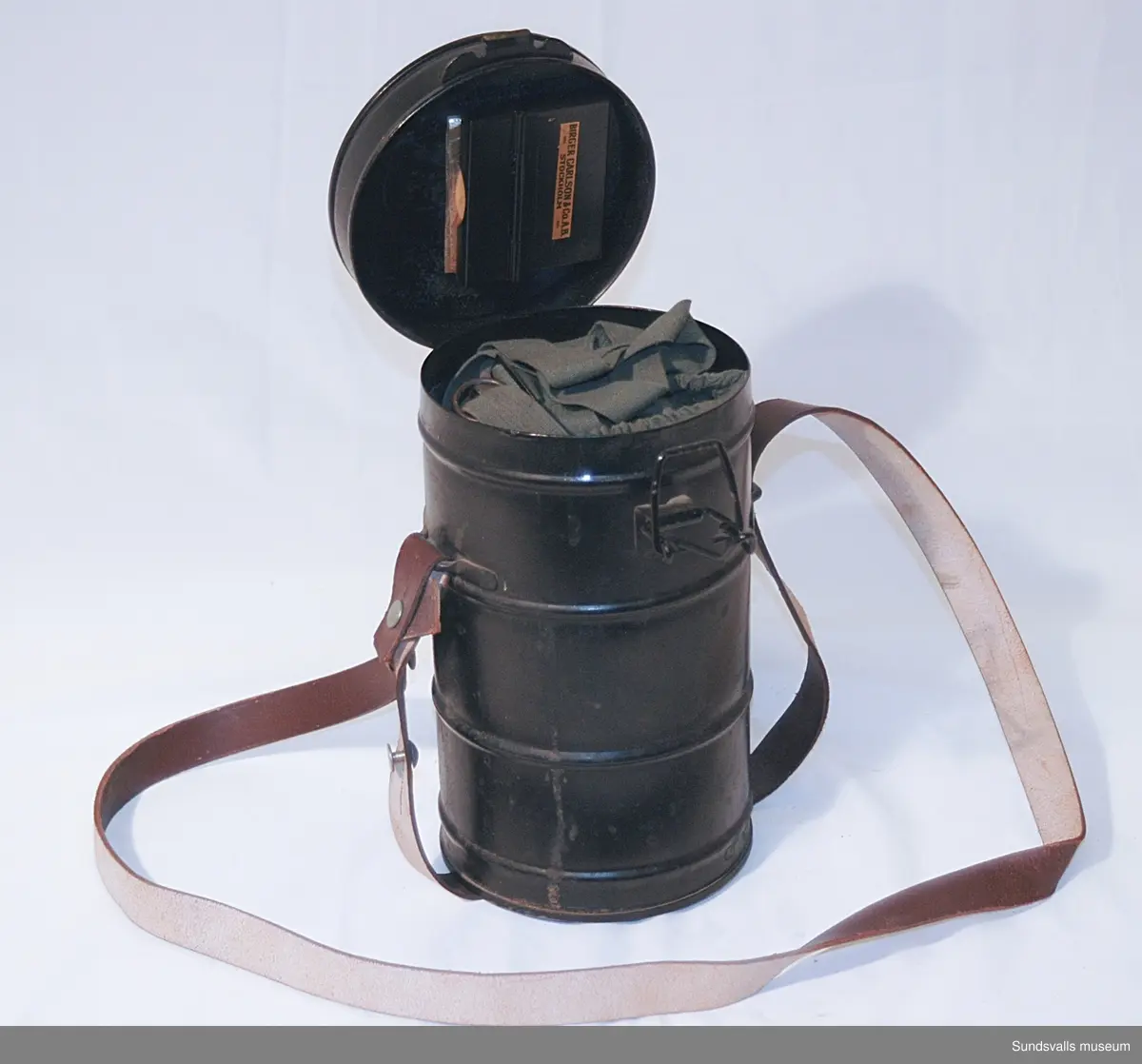 Gasmask/skyddsmask för ansiktet med filter mot föroreningar. Modellnamn 'Degea'. Förvaras i svart cylinderformad plåtbehållare med axelrem i brunt läder. I behållarens lock finns ett fack i vilket två sk. klarskivor förvaras i pappersomslag.  Masken är tillverkad av grönt bomullstyg i kraftig kvalitet, med runda metallinramade glas vid ögonen och en röd filterbehållare i metall. Invändigt är masken fodrad med sämskskinn. Masken spänns fast på huvudet med hjälp av resårband och reglerbara spännremmar.