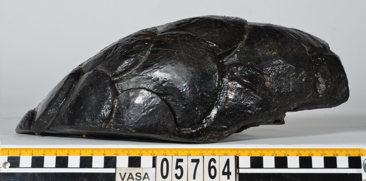 Del av skulptur, nedre del till fiskstjärt. Avsmalnande i ena änden. Rundad framsida med utsnidade fiskfjäll. Baksidan är bruten.