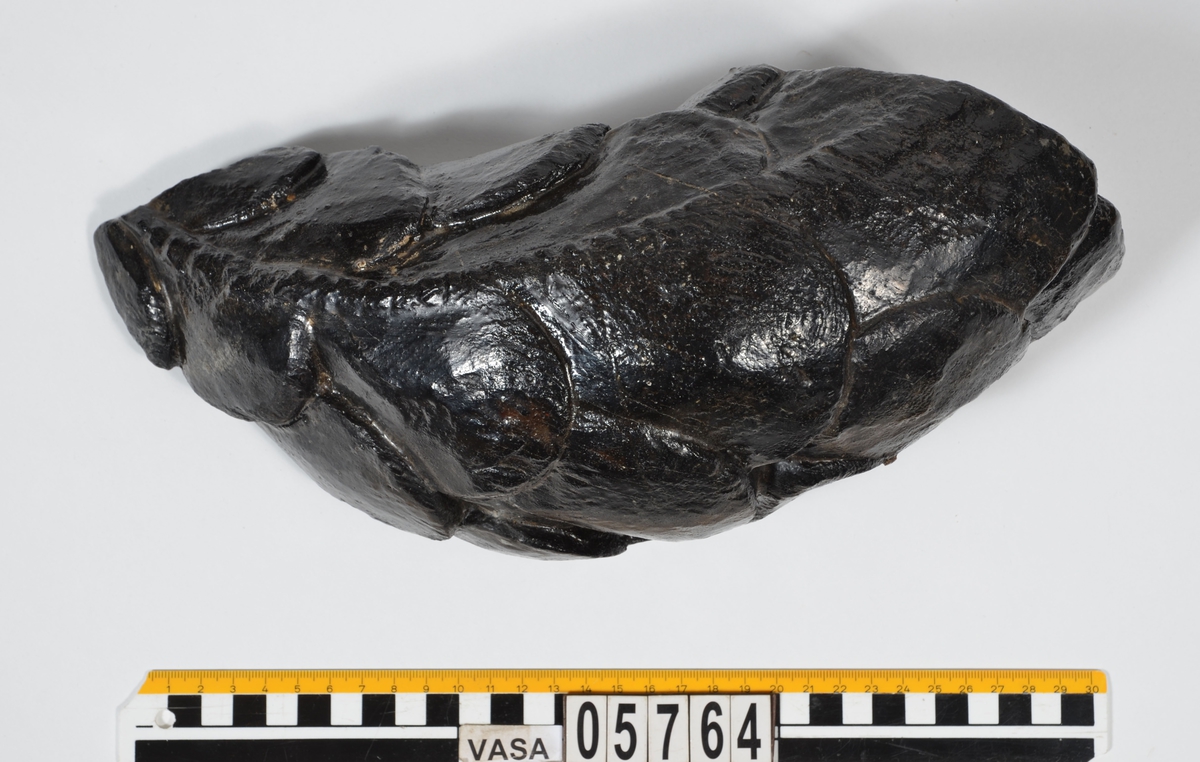 Del av skulptur, nedre del till fiskstjärt. Avsmalnande i ena änden. Rundad framsida med utsnidade fiskfjäll. Baksidan är bruten.