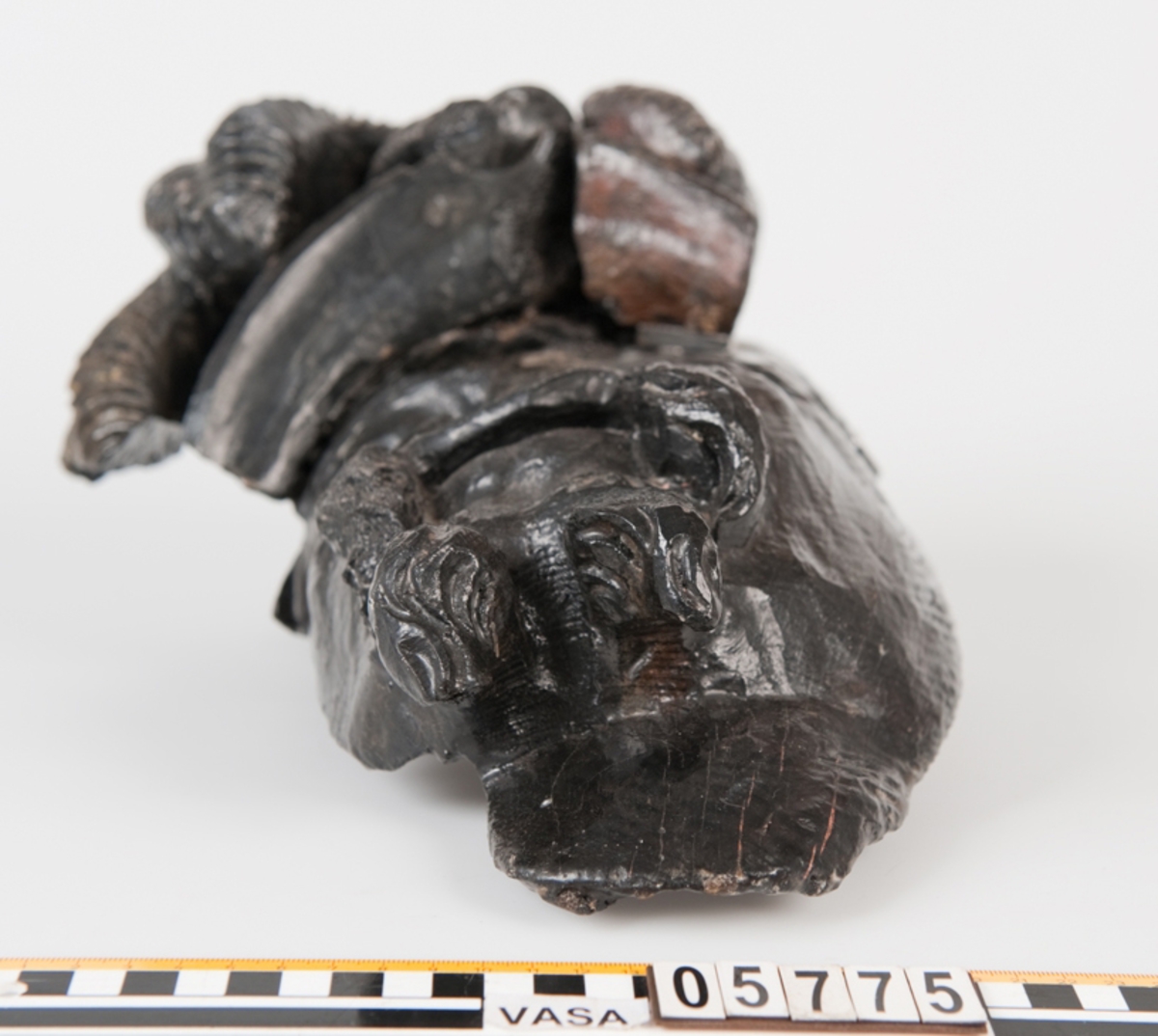 Del av skulptur. 
Huvudet till en högrest krigare med stor mustasch. På huvudet sitter hjälm med långa vajande plymer.