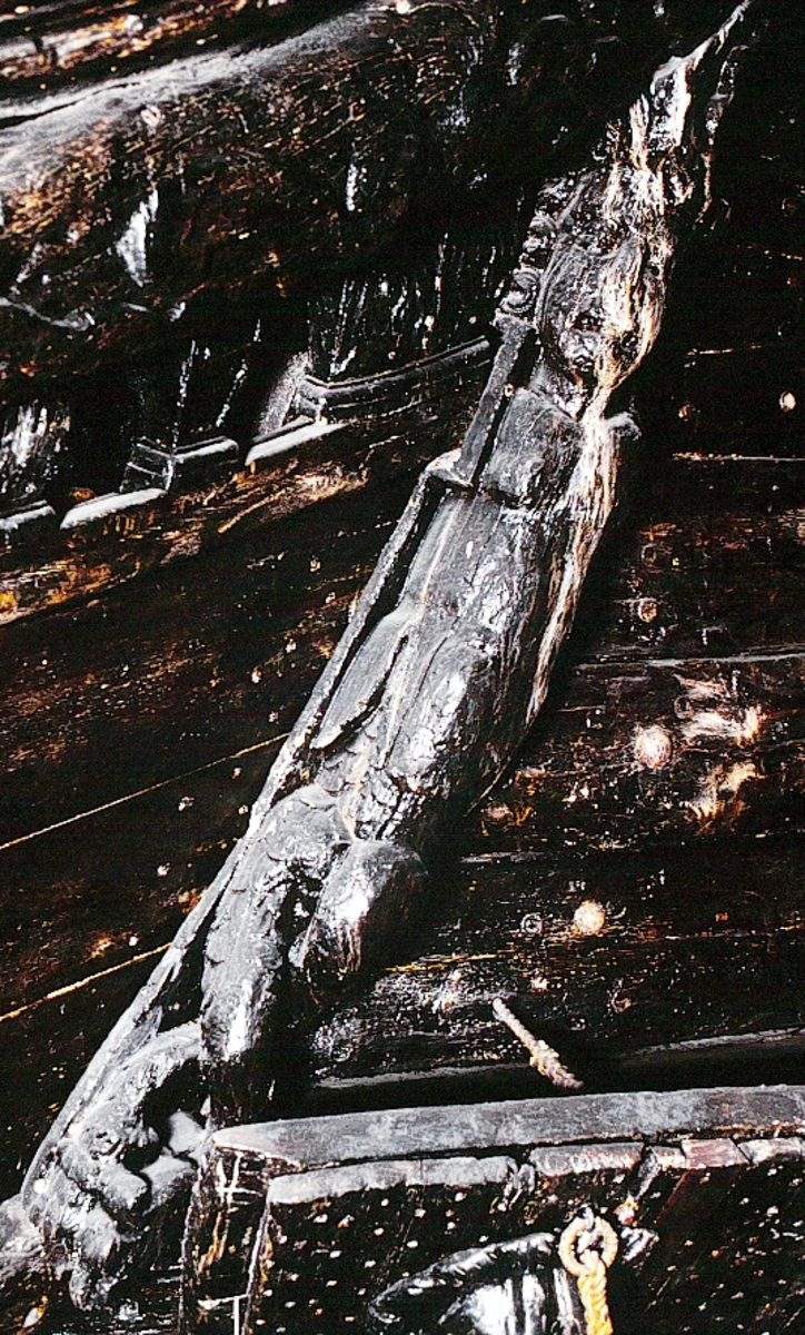 Stor, svängd triton med mansöverkropp och fiskstjärtar.

Skulpterad på tre sidor. Skulpturen är svängd mot vänster, sett från betraktaren. Tritonens ansikte och bål är vridna mot höger. Ansiktet är slätrakat och inramas av ringlande hårlockar. På hjässa bär tritonen en turbanliknande huvudbonad och vid vänster tinning ses ett stort snäckskal.



Armarna är ersatta av spiralliknande voluter. Överkroppen är täckt av en tunn, läderliknande klädnad som låter bålens muskler avteckna sig. Stora, bladlika flikar döljer övergången till underkroppens tudelade fiskstjärt.



Upptill är tritonen snett avslutad och dess högra sida, vänster sida sett från betraktaren, är utdragen i en spets. Vänstra sidan är avfasad och har utgjort anliggningsyta. Baksidan, som har utgjort anliggningsyta, har upptill ett snett, djupt och något avrundat urtag. Något längre ned, utmed höger kant, sett från betraktaren, återfinns en bred fasning.

Skulpturen är välbevarad, dock något sliten.



Text in English: A large curved Triton with male torso and fish-tails.

Sculpted on three sides. The sculpture curves to the observer''s left. The Triton''s face, which with the torso, is twisted to the right, is clean-shaven and framed by ringlets of hair. On the crown of the head is a turban-like headgear with a large sea shell on the left temple.



In place of arms, there are spirally rolled volutes. The upper part of the body is covered by a thin leather-like article of clothing that reveals the muscles of the torso, large leaf-like forms conceal the transition to the Triton''s lower parts which terminate in a double fish-tail.



Upwards, the Triton terminates crookedly, and its left side (the observer''s right) is extended into a point. Its left side is bevelled and has served as the attachment surface, along with the backformed as an attachment surface. The upper part of the back have a triangular cut-out hole and an obliquely placed cut-out hole that is somewhat rounded and conspicuously deep. Somewhat lower down there is a broad fastening.

The sculture is well preserved, but somewhat worn.