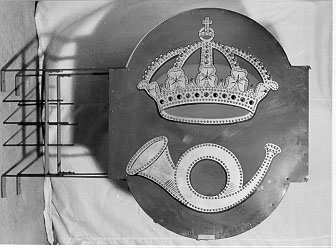 Postkontorsskylt av typ ljusskylt. Det krönta
postemblemetskonturer har hålats och fyllts med glaskulor i vitt,
rött och blått.Då skylten tänds tränger ljuset ut genom glaskulorna.
Skylten hartroligen varit uppsatt utanför postkontoret Stockholm 11
vidNytorget. Med metallställning för fastsättning på vägg.