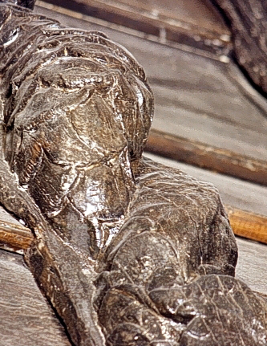 Triton med mustascher och tjockt, vågigt hår. Armarna är reducerade till upprullade bladflikar. Runt midjan sitter ett band med en oval medaljong i mitten. Skulpturens fiskstjärt slingrar sig kring stjärten till en fisk eller delfin, vars huvud är synligt vid skulpturens fotända.



Bakstycket är kraftigt och snidat i ett med skulpturen. Baksidan är försedd med hak eller urtag. Skulpturens profil är svagt S-formad.

Skulpturen är kraftigt sliten och anletsdragen i det närmaste utplånade.



Text in English: Triton with moustache and thick wavy hair. The arms are replaced by rolled-up leafy decoration. Round the waist there is a girdle or belt with oval decoration. The fish tail is entwined with the tail of a fish or dolphin whose head can be seen at the foot of the sculpture.



The backboard is robust and carved in one piece with the figure. The back side of the backboard was carved with hollowed out grooves. The profile is slightly S-shaped.

The sculpture is badly worn, the features have been practically obliterated.