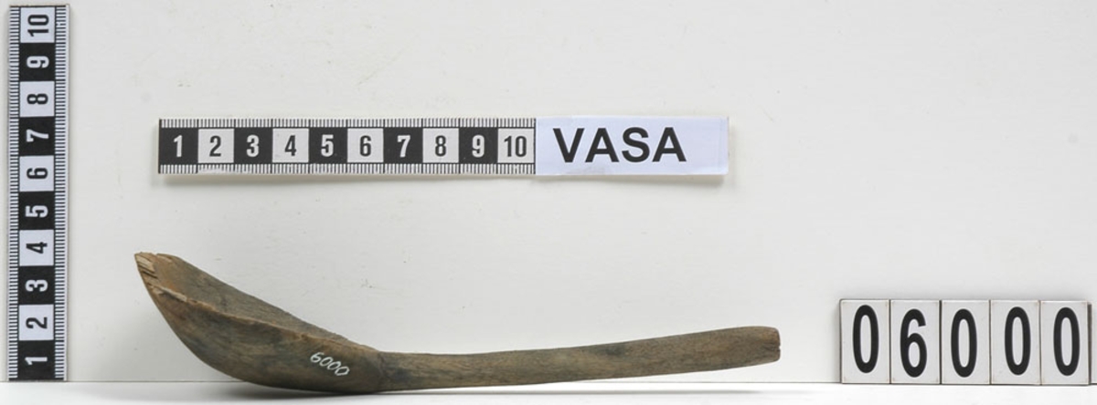En träsked där skedbladets kant är skadad. Skeden har en mörkbrun ton på baksidan vilket kan tyda på bemålning. Skaftet är ovalt.