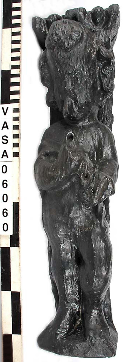 Skulptur föreställande en barnfigur som spelar på en sinka (ett äldre blåsinstrument med grepphål som blockflöjten men ett munstycke som liknar trumpetens).
Huvudet är vridet något åt höger. Kroppstyngden vilar på höger ben medan vänster ben är böjt. Instrumentet hålls med båda händerna. Skulpturen förefaller delvis draperad i en mantel eller dylikt som skymtar vid hans högra sida. Ovanför hjässan syns en något snedställd äggstavslist. Figuren står på ett minimalt underlag som troligen avser att föreställa ett stycke naturlig mark. Skulpturens baksidan är slät.
Skulpturen är kraftigt sliten.

Text in English: A sculpture of a child playing a sinka.
The weight of the body rests on the right leg while the left is bent and the head is turned slightly to the right.. The instrument is held in both hands. The figure seems to be partly draped in a cloak or the like, which conceals his right side. Just above the head the back board comes forward at right angles and the front of the board is decorated with an egg  mould design. The sculpture stands on a small flat plinth. The back of the sculpture is smooth.
The sculpture is very worn.