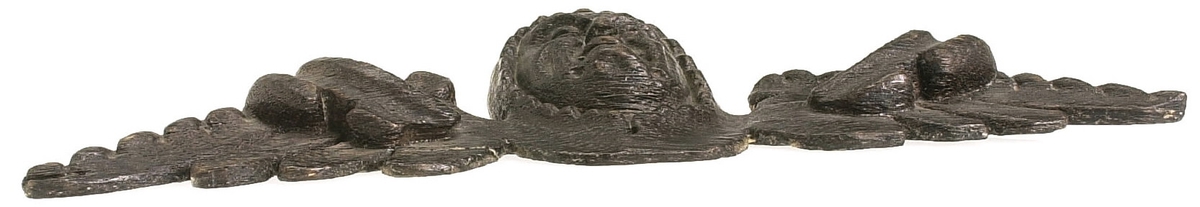 Kerub.
Skulptur i form av ett litet, runt kerub- eller gummansikte med böjd näsa, liten mun, tjocka kinder och kraftigt rynkad panna.
Ansiktet inramas helt av en tättslutande, mössliknande huvudbonad med flikig framkant. Den kombineras med en bred krage från vilken det utgår dekorativt formade vingar med pålagda broskornament. Baksidan är slät.
Skulpturen är mycket välbevarad.

Text in English: Sculpture of a small, round face of an old woman or cherub, with a curved nose, small mouth, plump cheeks, and heavily wrinkled forehead.
The face is wholly framed by a close-fitting covering like a night cap with a lobed front edge. This is combined with a wide collar from which extend large decoratively formed wings with applied earshell work. The back is flat.
The sculpture is very well preserved.