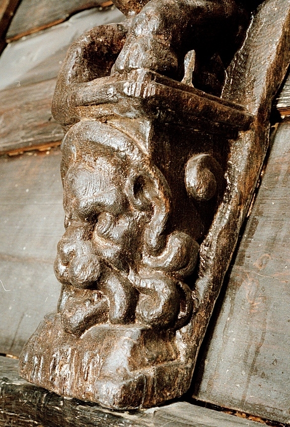Skulptur föreställande en djävulsfigur i starkt hukande ställning, placerad på en hög sockel.
Figurens huvud är utrustat med stora, krumma horn. I vardera handen håller den på ömse sidor av huvudet något som möjligen kan tolkas som en stor snäcka eller lur. Hakan pryds av ett bockskägg. Figuren har fladdermusvingar på ryggen och starkt luden underkropp med klövförsedda bockfötter. Fötterna vilar på den starkt sluttande sockeln, som framtill är prydd med en ap- eller lejonliknande maskaron med stor nos och hög, välvd panna. På sockelns sidor ses voluter.
Sockeln utgör fortsättning på det kraftiga klink-konstruerade bakstycket som är framdraget ovanför figuren.
Skulpturen är delvis kraftigt sliten och vittrad, särskilt den övre delen. Nederdelen är något bättre bevarad.

Text in English: Sculpture of a devil figure in a crouched position, placed on a high socle.
The figure''s head is equipped with large, curved horns. In either hand the figure holds on either side of its head something that may possibly be identified as a large seashell or horn. The devil has a goat''s beard on its chin, bat wings on its back and very hirsute lower parts of the body, and cloven goats'' feet.
The feet rests on a sharply sloping socle that is decked in front with a mask of an ape-like or lion-like face with a large nose and a high arched forehead. On the sides of the socle volutes are visible.
The socle forms the extension of the heavy, clinker-built back-piece, which is drawn forward above the figure.
The sculpture, particularly its upper parts, is much worn and disintegrated. The lower part is somewhat better preserved.
