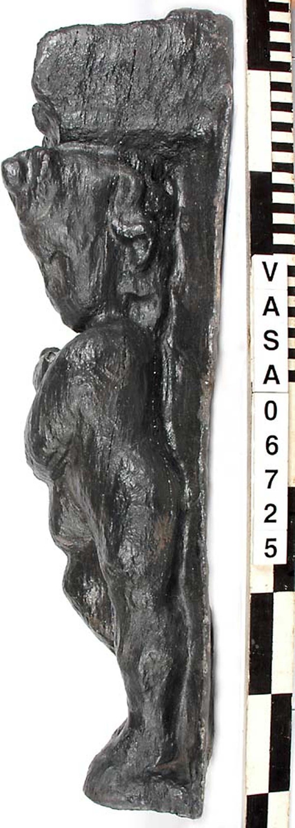 Skulptur föreställande en barnfigur spelande på en säckpipa.
Huvudet är vridet åt vänster och något nerböjt. Säckpipan hålls med bägge händerna och stöder dessutom på höger ben som är böjt. Kroppstyngden vilar på vänster ben.
Ovanför hjässan ses en snedställd äggstavslist. Det triangelformade partiet ovanför listen har utgjort anliggningsyta. Figuren står på ett minimalt underlag som troligen avser att föreställa ett stycke naturlig mark. Skulpturen har en slät baksida.
Skulpturen är kraftigt sliten och ytnött.

Text in English: This is a sculpture of a child playing a set of bagpipes.
The bagpipes are held in both hands and are further supported on the right leg, which is bent. The left leg is straight and takes the weight of the body. The head is turned slightly to the right and a trifle bent. The figure is partly draped in a cloak which conceals his right side.
Above the head the backboard comes forward at right angles. The front of the board is wide and is decorated with an egg-moulding design  which is placed diagonally across it, with the left-hand side lower than the right hand side when viewed from the front.. The figure stands upon a small flat plinth. The back of the sculpture is smooth.
The sculpture is badly worn.