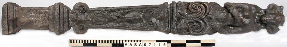 Skulpterad pilaster med övre delen i form av en karyatid med yppig, konstrik frisyr.
Höger arm hålls böjd med handen placerad framför bröstet. Vänster arm hålls utsträckt utefter sidan med handen vilande mot höften. Underkroppen döljs av ett hölje som framtill är prytt med stora, spiralformade ornament och små djurfigurer.
Karyatiden avgränsas från det rullverksornerade pilasterskaftet av en profilerad list. Nedtill avslutas skaftet av ett rullverksornament ovanför den rikt profilerade basen. Nedtill är pilastern försedd med en hög piedestal, prydd med beslagsornamentik. Figuren och dekoren omfattar även pilasterns sidor. Baksidan är slät.


Text in English: Sculpted pilaster with its upper part shaped as a Caryatid with a luxuriant artistic coiffure.
The right arm is held bent with the hand in front of the breast, the left arm is held at the side with the hand resting against the thigh. The lower part of the body is concealed by a shift or slip that is decorated with large spiral-shaped ornaments and small figures of animals.
A profiled batten separates the Caryatid from the shaft of the pilaster, which is ornamented with scrollwork. The shaft terminates with a scrollwork ornament above the richly profiled base. Below, the pilster is equipped with a high pedestal, decked with applied ornamentation. The back is smooth.