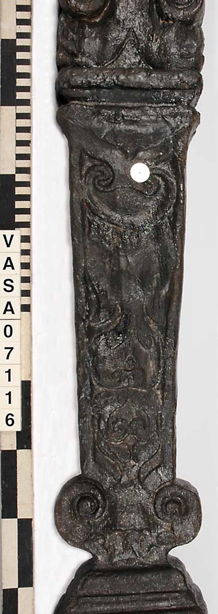 Skulpterad pilaster med övre delen i form av en karyatid med yppig, konstrik frisyr.
Höger arm hålls böjd med handen placerad framför bröstet. Vänster arm hålls utsträckt utefter sidan med handen vilande mot höften. Underkroppen döljs av ett hölje som framtill är prytt med stora, spiralformade ornament och små djurfigurer.
Karyatiden avgränsas från det rullverksornerade pilasterskaftet av en profilerad list. Nedtill avslutas skaftet av ett rullverksornament ovanför den rikt profilerade basen. Nedtill är pilastern försedd med en hög piedestal, prydd med beslagsornamentik. Figuren och dekoren omfattar även pilasterns sidor. Baksidan är slät.


Text in English: Sculpted pilaster with its upper part shaped as a Caryatid with a luxuriant artistic coiffure.
The right arm is held bent with the hand in front of the breast, the left arm is held at the side with the hand resting against the thigh. The lower part of the body is concealed by a shift or slip that is decorated with large spiral-shaped ornaments and small figures of animals.
A profiled batten separates the Caryatid from the shaft of the pilaster, which is ornamented with scrollwork. The shaft terminates with a scrollwork ornament above the richly profiled base. Below, the pilster is equipped with a high pedestal, decked with applied ornamentation. The back is smooth.