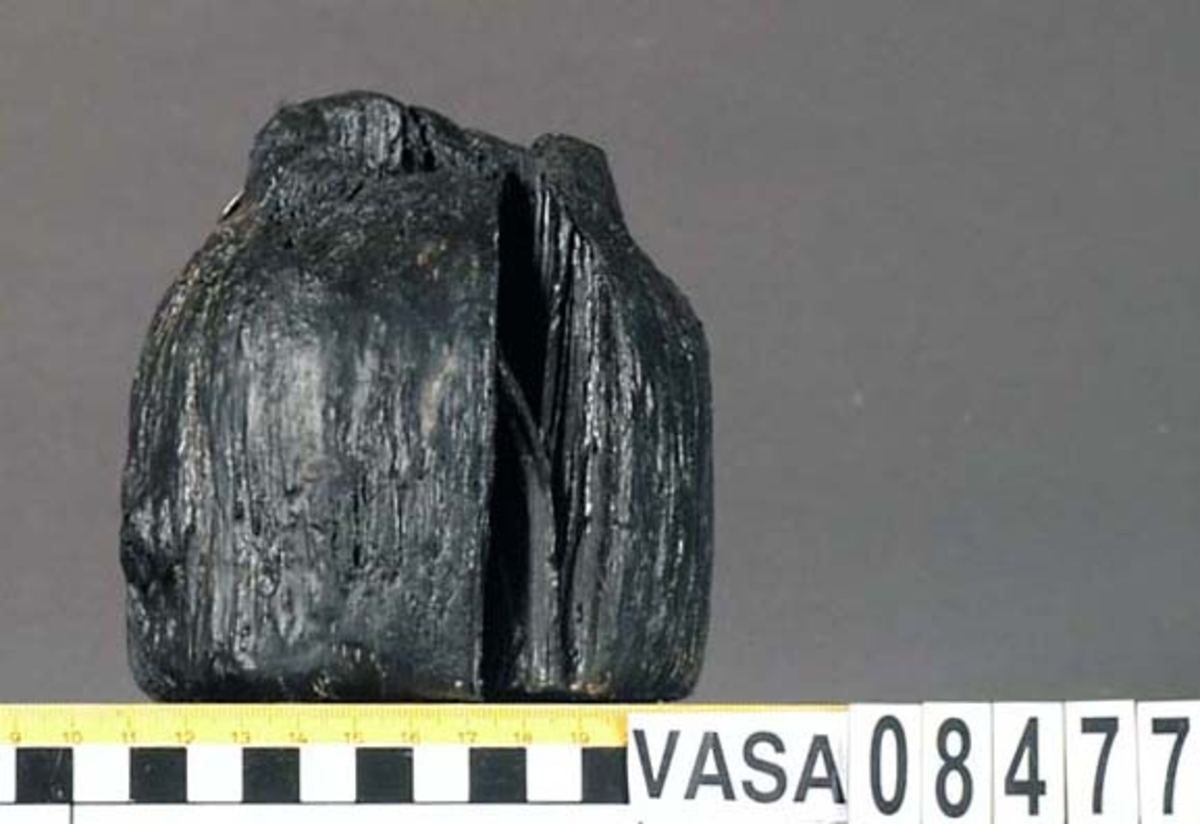 Kolv till ansättare. Kolven består av ett cylindriskt trästycke med ett centrerat genomgående skafthål. I övre änden är kolven profilsvarvad medan den nedre änden avslutas tvärt. Kraftigt eroderad, inga spår av svarvningen. En kraftig spricka samt ett antal mindre.