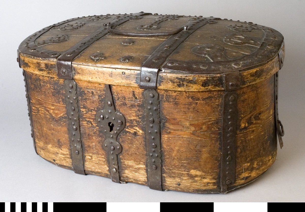 Färdskrin av trä med beslag av järn. Oval form med ett lock som är dekorerat med smida järnbeslag. Kanten på locket är skod med järn. På framsidan löper beslag ned och runt om. Centrerat mitt fram finns ett lås med nyckelskylt i järn.