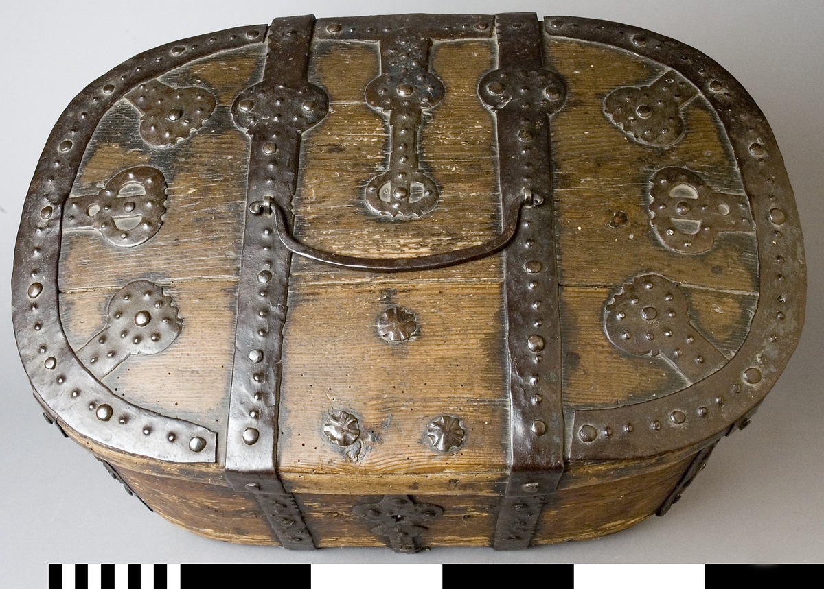 Färdskrin av trä med beslag av järn. Oval form med ett lock som är dekorerat med smida järnbeslag. Kanten på locket är skod med järn. På framsidan löper beslag ned och runt om. Centrerat mitt fram finns ett lås med nyckelskylt i järn.