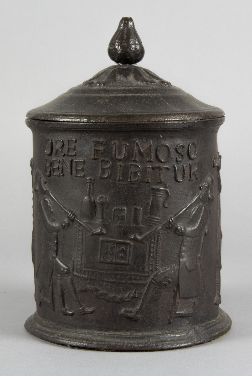 Relief på burken med text. Relief med rökande och spelande män. Några av männen sitter vid en spis. Riksvapen med flankerande lejon. Årtal 1794.
Text på burken. ORE FUMOSO BENE BIBENTUR.