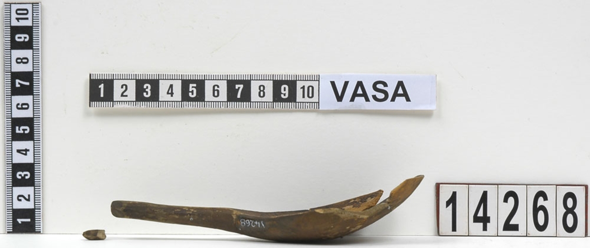 En träsked, ursprungligen bruten i sex delar. Den största delen utgjordes av skedskaftet samt en del av skedbladet. Fyra av delarna tillhörde skedbladet. Den minsta delen utgjordes av en liten knopp, vilken suttit i skedskaftets ände. Skeden är idag sammanfogad av konserveringsenheten.
Skedbladet är äggformat och relativt stort i jämförelse med skaftet. På insida av skedbladet - längs med ytterkanterna, intill skaftet - syns en inristad dekor i form av linjer och streck. Skaftet är kort och rakt, med profilerad ände. På skedbladets baksida, intill skaftet, är ett bomärke inristat. På skeden syns rikliga spår av rödbrun färg.
Skeden är mycket ömtålig. Skedbladets ytterkanter saknas delvis.