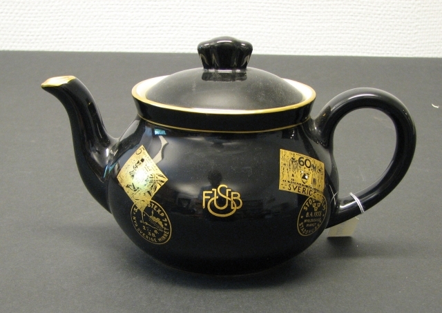 Tekanna i svart porslin med frimärksmotiv och stämpelavtryck
i guldfärg:  Nya Sverige Minnet 5 öre, 2 x 60 öre och 20 öre, 1938,
stämpelavtryck med text: Stockholm Nya Sverige minnet Stadshuset;
Sjöpostexp 7 Nya Sverige minnet.