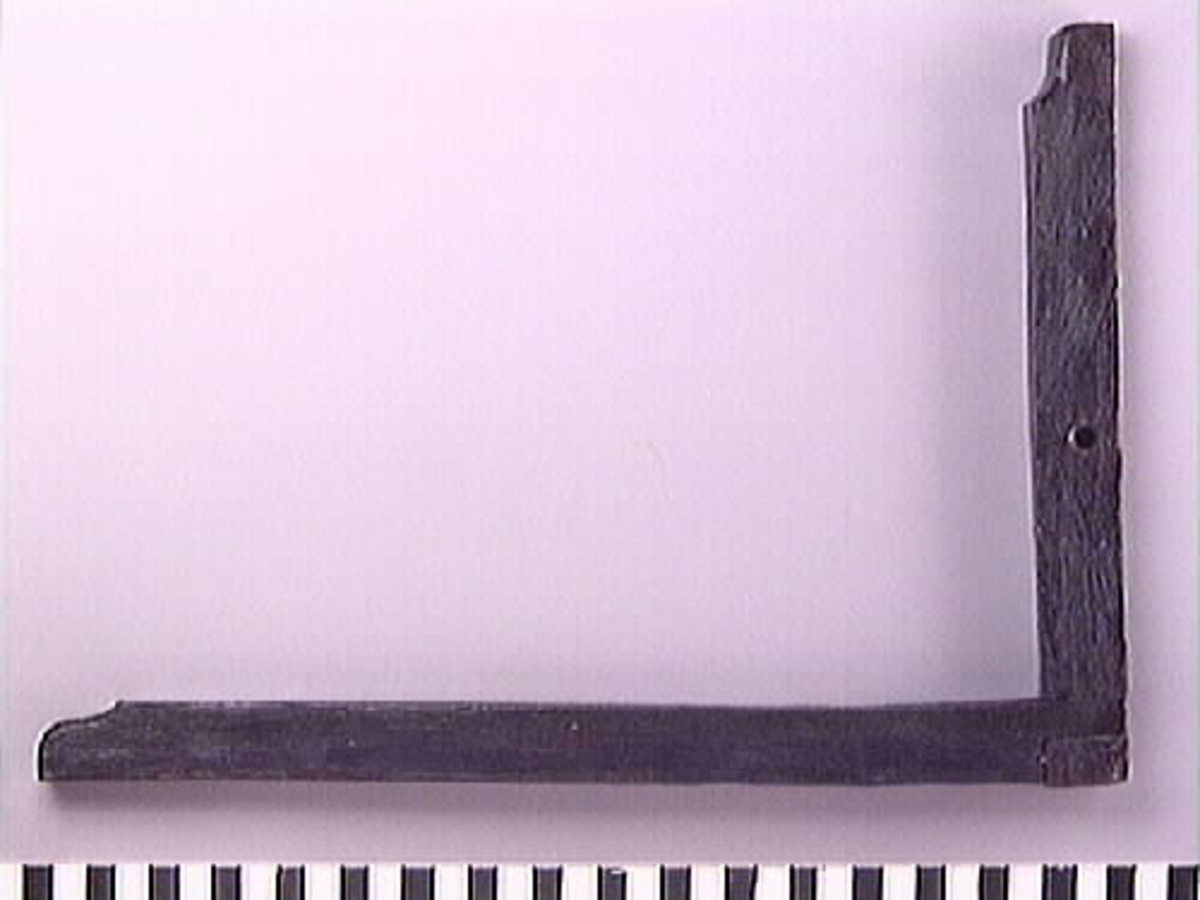 En vinkelhake.

Vinkelhaken består av två plana stavar som är sammanfogade genom sinkning. Ändarna är profilerade. I mitten av den kortare staven finns ett runt hål för upphängning.

Vinkelhaken är välbevarad och i gott skick.

För mer information se Dokumentationsblankett under Referenser – Länkade filer.