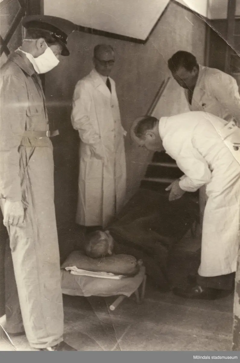 "Karantänsförläggning" på Kvarnbyskolan för överlevande från koncentrationsläger i Tyskland och Polen 1945.  
Skolan fungerade under denna sommar som beredsskapssjukhus.

En överlevande ligger på en bår, omgiven av tre läkare och en Röda Kors-arbetare.