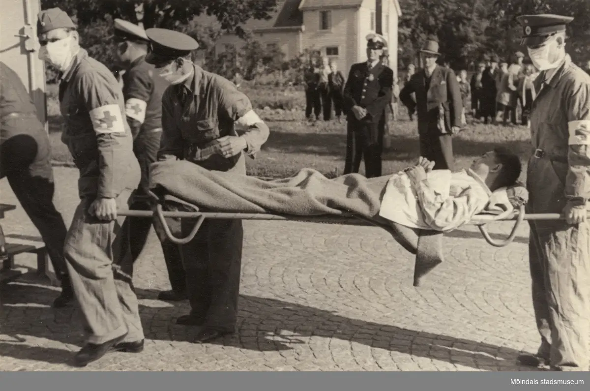 "Karantänsförläggning" på Kvarnbyskolan för överlevande från koncentrationsläger i Tyskland och Polen 1945.  
Skolan fungerade under denna sommar som beredsskapssjukhus.

En manlig flykting på bår, buren av Röda Korsets personal. I bakgrunden syns polis och nyfikna åskådare.