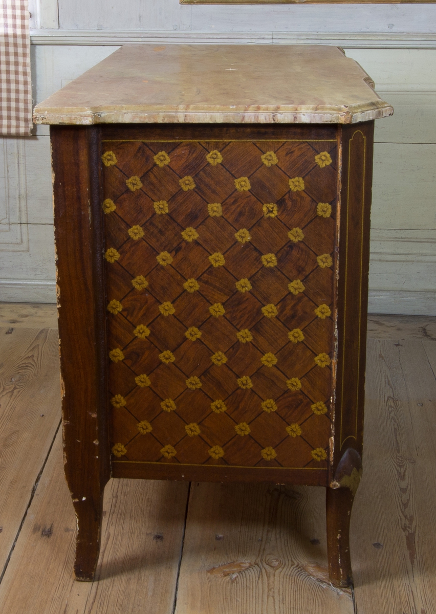 Byrå av furu, med imitationsmåleri som efterliknar fanér och inläggningar i olika träslag, i form av rutinläggningar och blommor. Med marmorerad skiva av trä. Rak form med avfasade hörn, med svagt svängda ben. Fronten med förkroppat mittparti, som nedåt avslutas i svagt bågformat undergehäng. Med tre lådor med nyckelbrickor med bandrosetter och beslag med dragringar i form av lagerkransar av förgylld mässing.