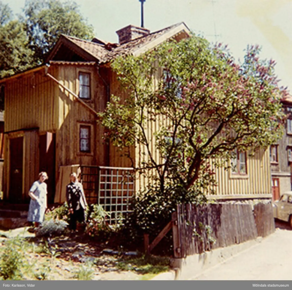 Bostadshuset Roten F25, Trädgården, år 1968. Ena gaveln med ett träd och några personer vid sidan av.