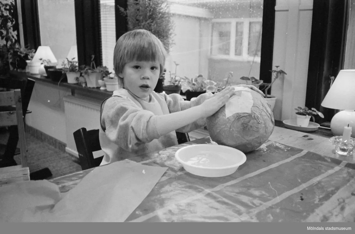 En pojke sitter vid ett bord, inomhus på Katrinebergs daghem, och limmar silkespapper på en uppblåst ballong. Limmet ligger på en plasttallrik framför honom och han tittar mot kameran.