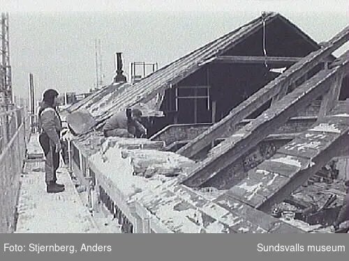 Bild 05 Rivning av takstolar norra mittpartiet. Taket med falsad plåt och undetaket  av bräder är redan bitvis borta.Bild 06 Detalj av stålbalk med stöd mot yttervägg i norr.Bild 08-09 Överbyggnaden inifrån mot sydöst.Bild 16,19 Vy mot frilagd skorsten i söder dvs den dubbla skorstenens högra del.Bild 20-21 Byggnadens norra del fr V.Bild 22 Den bortplockade vapensköldens väggyta på norra fasadens risalit.Bild 23 Vy utmed norra takfallet fr V