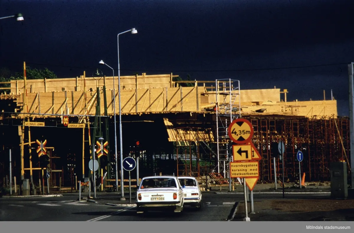Mölndals Bro under uppförande, år 1975. Framför bygget står ett par bilar på ett övergångsställe. Vid sidan av ses trafikskyltar.