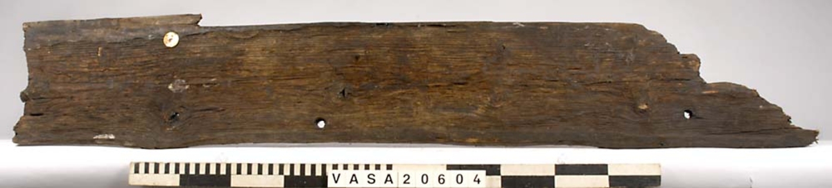 Bordläggningsbräda till skeppsbåt.
Alla kanter är avbrutna utom en liten kant. 2 plugghål/dymlingshål.

Text in English: straight pine board. Upper outside edge is beveled. Lower edge is beveled with luting cove.
Few iron nails, no head impressions. Two diameter 15mm treenail holes, champfer on the outboard face.
surface erosion on the outboard face. Inboard face is better preserved. Breakage, only small pieces of the original edge exist. Ghosts on both outside edges