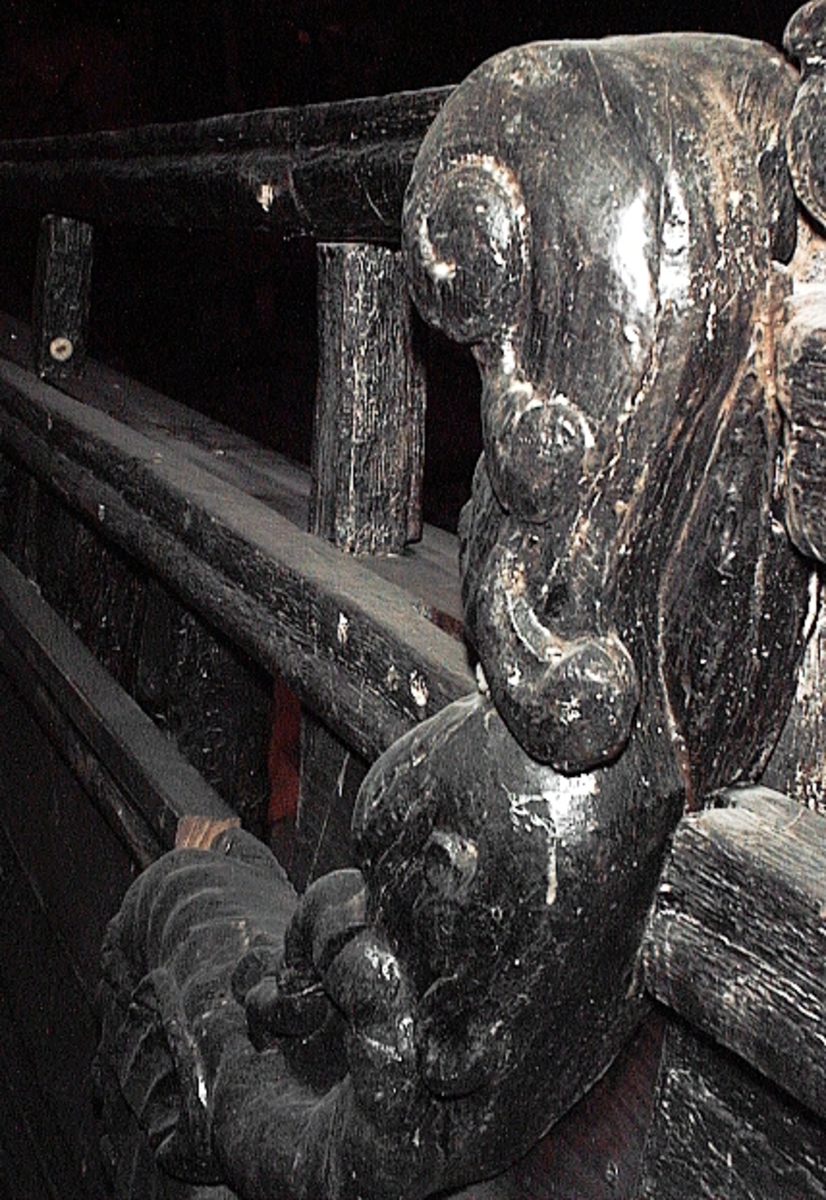 Skulptur av en triton, svängd mot höger.
Tritonen är framställd i vänster profil och avtecknar sig i hög relief mot det släta bakstycket. Tritonen har en stor näsa och treflikig mustasch. På huvudet sitter en hjälm med dekorativa bladvoluter. Armarna är ersatta av långa, flikiga bladslingor. Bålens övergång i den fjällförsedda fiskstjärten markeras av stora bladbildningar. Tritonens stjärt döljs bakom stjärten och vingen till en under liggande drake, se fyndnummer 21277. På baksidan finns urtag för relingslisten.
Skulpturen är mycket välbevarad.

Text in English: A sculpture of a Triton curved to the right.
The figure is seen in left profile, in high relief against a plain back board. The face has a large nose and a moustache that splits into three parts. On its head is a helmet with decorative leaf volutes. The arms are replaced by long, lobed leaf formations. The transition area between the male torso and the scaled fish-tail is decorated with large leaves. The rear is hidden behind a dragon''s coiled tail and broad wing situated below, see No. 21277. There are notches on the back that fit into the railing.
The sculpture is very well preserved.