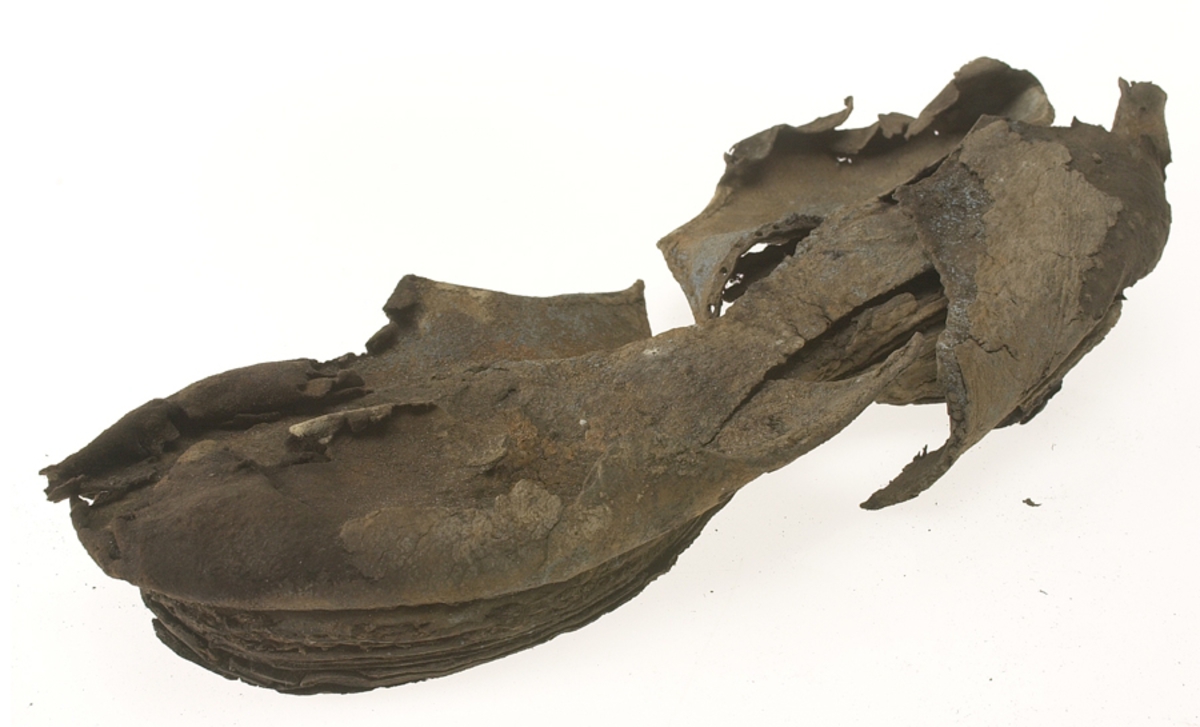 Sko.
Inuti skorna hittades även fragment från olika textilier som ursprungligen varit ett par strumpor. Idag sitter ett stickat fragment av ull fast i vänsterskon (23070) med avigsidan uppåt. I fyndet finns även en mängd lösa stickade fragment som idag är mycket korroderade och vissa helt mineraliserade av järn vilket har gjort textilen stenhård och därmed oerhört skör (23070a-c). Utifrån minskningar som några små fragment har så har det varit möjligt att rekonstruera strumpans tå. Genom att pussla ihop fragmenten som ursprungligen har suttit ihop har det på så vis gått att avgöra hur strumpans tå har varit konstruerad. Då textilen är så fragmentarisk är det inte möjligt att avgöra hur resten av strumpan har sett ut eller varit konstruerad. Däremot så finns det fragment av ett kypertvävt tyg av ull som på vissa ställen ligger dubbelt med det stickade 23070c). Ett av dessa fragment har stelnat i formen så som den suttit på fotens främre del inuti skon. Detta fragment täcker delvis det stickade tyget och har dessutom spår av kaststygn längs en kant. Så som det vävda tyget är placerat på det stickade tyget pekar på att man använt det som lagningslappar till sina stickade strumpor. 
I fyndet finns det även korroderade järnklumpar, ett tuskaftsvävt tyg av ull och rester av ett grovt bastfibergarn och två mindre läderfragment (23070d-f). Det är möjligt att även dessa har tillhört strumpan och/eller skon.

För mer detaljerad information från textildokumentationen (år 2022) se länkade filer.

Skodokumentation 2022:
Ovanläder. Tillverkad i getskinn av god kvalitet. Hör ihop med fyndnummer 23069 (Yngves skor). Tillverkades på en rak läst och bars på vänster fot. Inga dekorationer Det finns stygnhål som visar att det funnits invändiga förstärkningar (men förstärkningslappen saknas). Det finns delaminering men inga lagningar.
För mer information, se Länkade filer: Uppritning

Text in english:
This is the upper of a shoe of more “extreme fashion”, made of goatskin leather of good quality, probably made on a straight last and worn of the left foot, with no decorative features. The upper couples with 23069, together composing Yngve’s shoes. There are internal reinforcements missing from the upper and delamination, there are no repairs.
For more information, go to Länkade filer: Uppritning