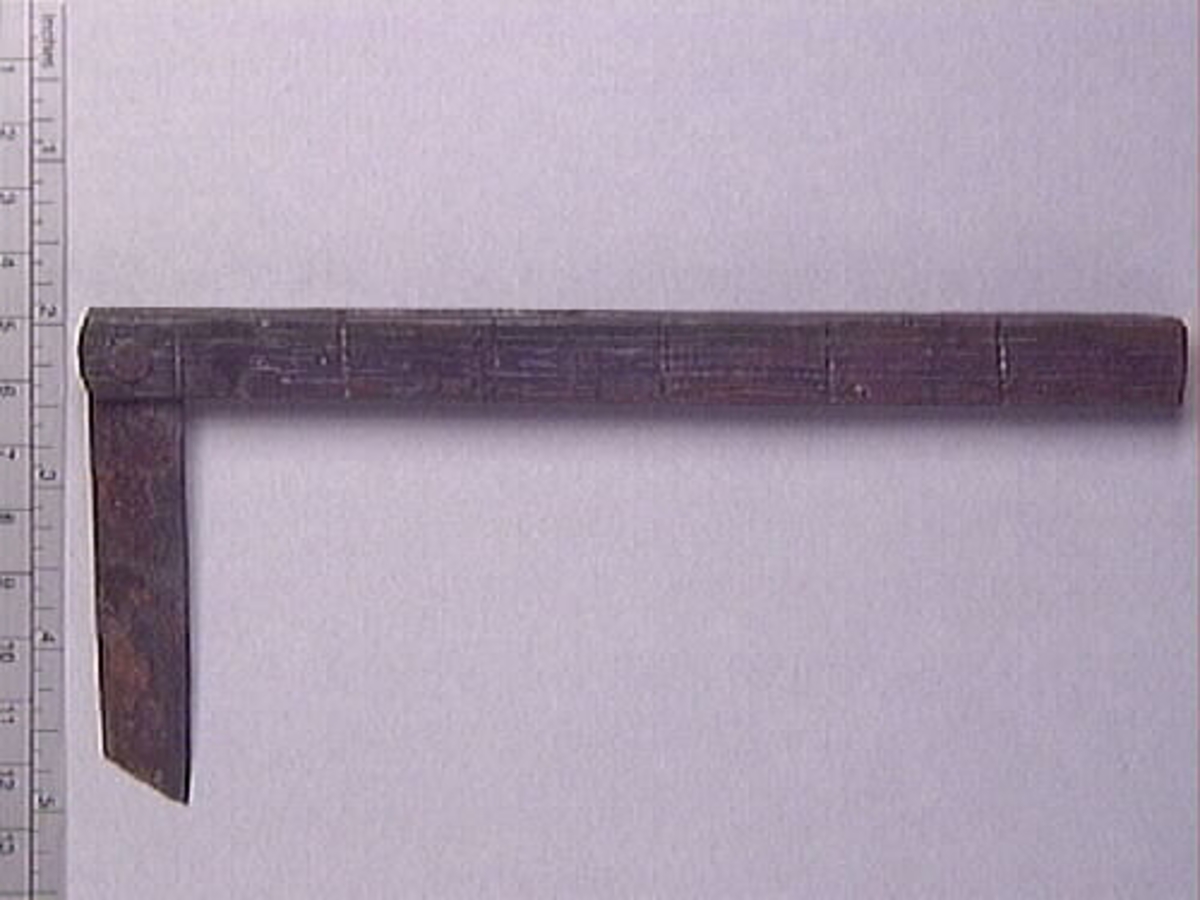 En vinkelhake, s k smygvinkel.

Skaftet är försett med sex stycken ristade skåror som markerar tum. I ena skaftänden sitter ett nytillverkat, ledat, järnblad. Bladet är fäst med en nytillverkad järnsplint. I skaftets ena ände, närmast bladet, löper en skåra eller ett spår. Spåret är mellan 68 och 80 mm djupt.

Smygvinkeln är i gott skick.

För mer information se Dokumentationsblankett under Referenser – Länkade filer.