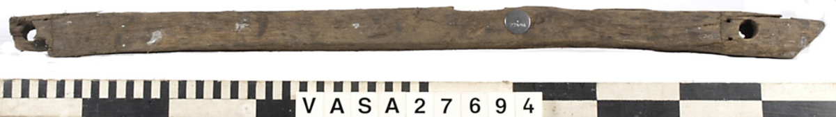 Relingslist. Den ena kortänden är uppfläkt. Den andra kortänden avslutas med en snedfasning, sannolikt en lask. Den andra slutar tvärt i sambands med en ursparning (sannolikt platsen för en årtull). Tolv stycken ej genomgående spikar i vad som sannolikt är den sida som vetter mot bordläggningen indikerar att den har spikats fast utifrån. Ett kraftigt genomgående hål syns i anslutning till ursparningarna för årtullarna. Spikhål syns i dessa avtrycks hörnor. På den sidan som vänder mot bordsidan och på undersidan syns snedfasade urtag, sannolikt för topparna till intimmer. I dessa urtagningar kan man skönja tydliga verktygsspår.