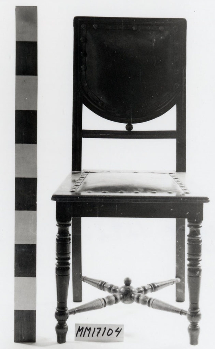 Stol, troligen av ek, fernissad och betsad i mahogny. Frambenen är raka, runda och svarvade, bakbenen är fyrkantiga och svagt utåtsvängda. I kryss mellan stolsbenen sitter svarvade tvärslåar, lågt placerade. De förenas mitt under stolen i en träkula med en mindre kula ovanpå. Bakbenen fortsätter i ryggens sidostolpar som utgör kanterna av ryggbrickan, rak i överkant och sidor, halvcirkelformad i nederkanten. Ryggbrickans träram är profilerad på framsidan och i mitten klädd med brunt läder, fästat med stora läderklädda nitar. Ryggbrickan vilar på en mindre träkula som pressas mellan denna och tvärslån under. Sitsen har raka sargar, sidosargarna är rakt vinklade framåt, profilerade kanter. I mitten klädsel av brunt skinn, fästat med lädernitar. Stoppning invändigt, samt undertill klädd med fanér. Märkt: "TPHN" samt så kallad kattfot.