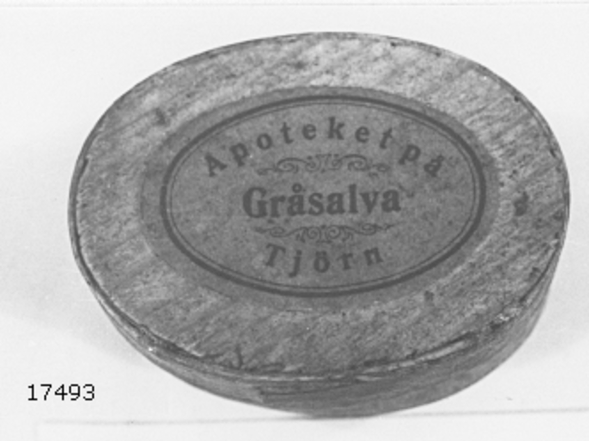 Ask av furuspån, liknande svepask, oval form försedd med lock. På lockets ovansida oval etikett, rosa med svart text: "Gråsalva Apoteket på Tjörn" samt tunna dekorslingor. Invändigt rester av salvan.