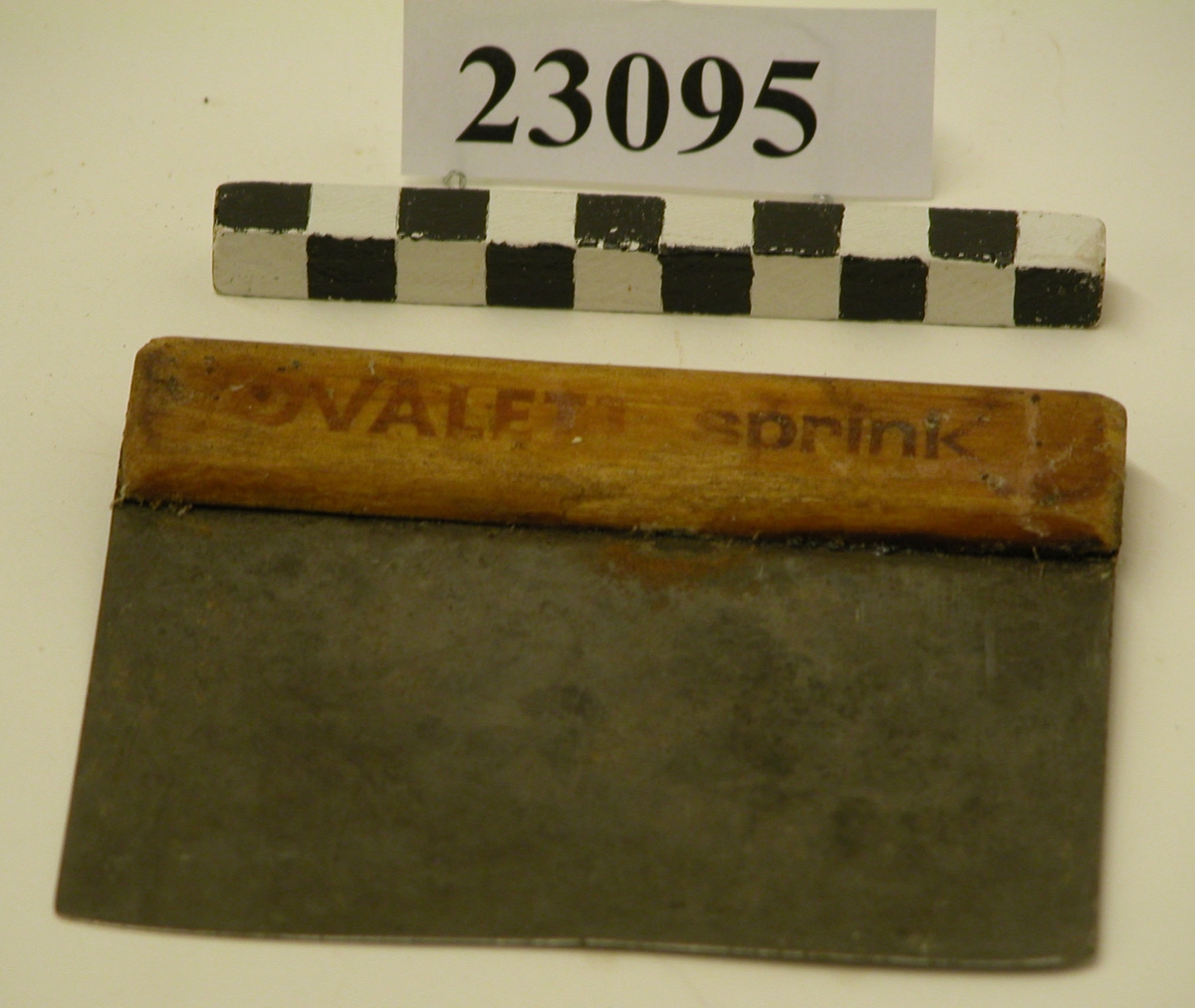 Degskrapa av tunn järnplåt försedd med handtag av trä. Plåten instucken i spår i träet och fäst med två stift av metall.  På handtaget står med rödbruna bokstäver tryckt texten "OVALETT sprink".