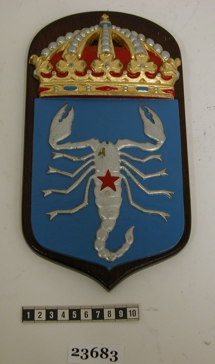 Vapensköld typ B, torpedbåten Antares. Vapensköld med fullständig kunglig krona på bottenplatta av teak. Blåfärgad sköld. På skölden finns en silverfärgad skorpion försedd med en röd stjärna på ryggen. Bottenplattan är på baksidan märkt med nummer 43, skrivet med svart tusch för hand.