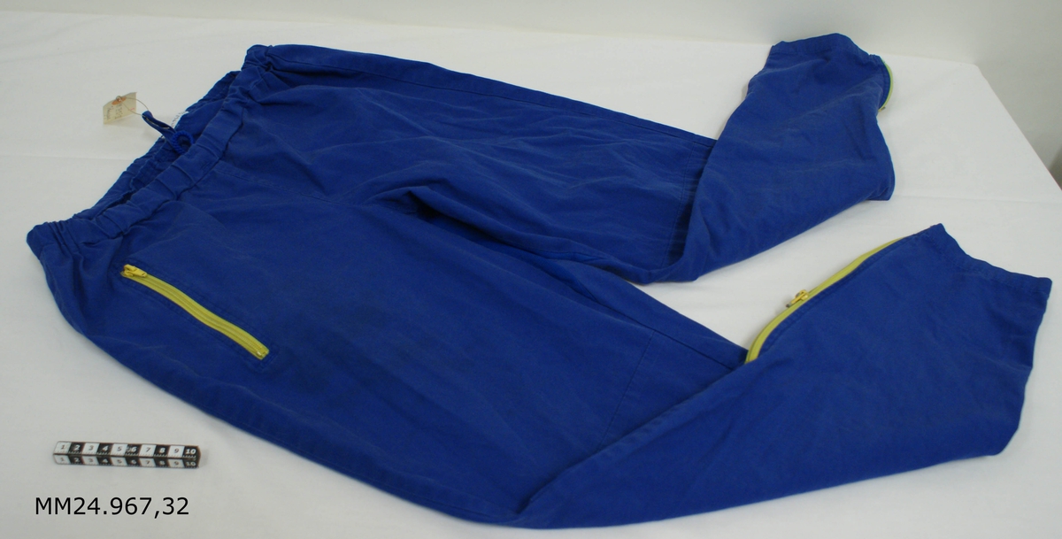 Träningsbyxa av poplintyg häften bomull hälften polyester. Klarblå byxa med gult blixtlås på sidofickan samt vid byxbenens slut. Resår och snörning i midjan. Annat tyg i skrevet för bättre luftcirkulation. Vitlapp på insidan med kronstämpel, M-nummer, tvättråd, årtalet 2005, vikt 70-80 kg, storlek 5, samt texten: "Suntex Group Co Ltd, Peoples Republic of China".