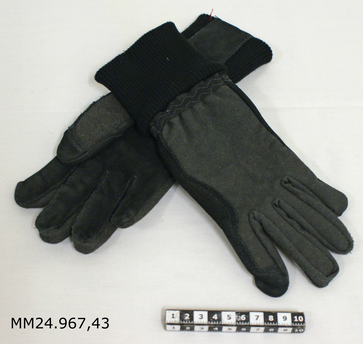 Svarta handskar med bomullsmuff och kevlarförstärkt ovandel. Femfingermodell. Inne i varje handske sitter en vit tyglapp med information om storlek, material med mera.