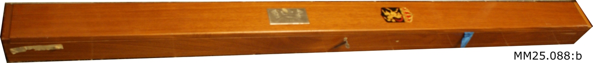 Rektangulär trälåda i mörkt fernissat trä. Lådan är försedd med lock och lås. På lådans lock finns en målad vapensköld med en get som står på bakbenen. Vapnet försett med krona. Längre ner sitter en metallskylt med två korsade flaggor, flankerade av två vapensköldar, samt text "Jagaren Hälsingland 1959" ingraverat.