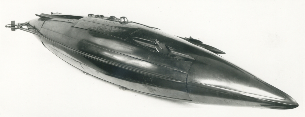 Spolformad. Stålplåt. Slingerköl, 2 propellrar av metall, 2 st höjd- och sidoroder parallellkopplade med gardjärn. 2 st höj- och sänkbara periskop av mässing. 4 st rännor för torpeder, därav 2 med luckor framtill, dels 2 för magasinering. Kort cylinder, rörlig i ränna midskepps akterut. Torpedrännorna täckas av långa stållock. Tillhörande 4 st torpeder av massiv mässing. Eneroths förslag till ubåt 1897. UndervattenstorpedFöremålets form: Spolformad.