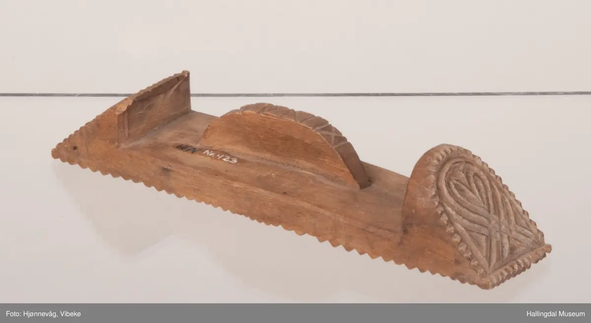 Rektangulær med skråttstilte flater i sidene. Mønsteret består i bunnen av to like felt, blomster dannet av to hjerter stilt med spissene mot hverandre. Tausnoingsborder langs kantene. Den ene sideflaten er nesten kvadratisk, hjertemønster omgitt av tausnoingsbord. Den andre flaten har en rett side nederst, oval øverst, hjertemønster og tausnoingsbord. Baksiden har håndtak, skåret i ett med stemplet. Buet med innskårne kryss på ryggen. Oppl. gml. prot.: smørknott.

23-7-1963. Å. Astrup.