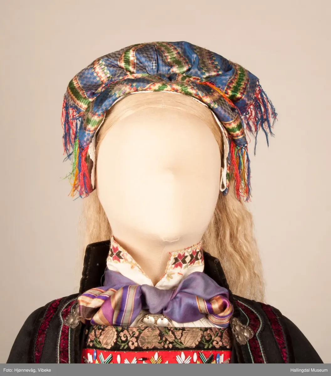 Kone fra Øvre Hallingdal i hallingbunad.  Hun bærer hette.  Kona står utstilt i den faste bunadutstillingen på Hallingdal Museum, Nesbyen.  Hallingbunaden består av mange draktdeler; trøye, Forkle HFN 02033, stakk, hette, silketørkle i halsen, perlet bringduk el. brystklut og skjorte.