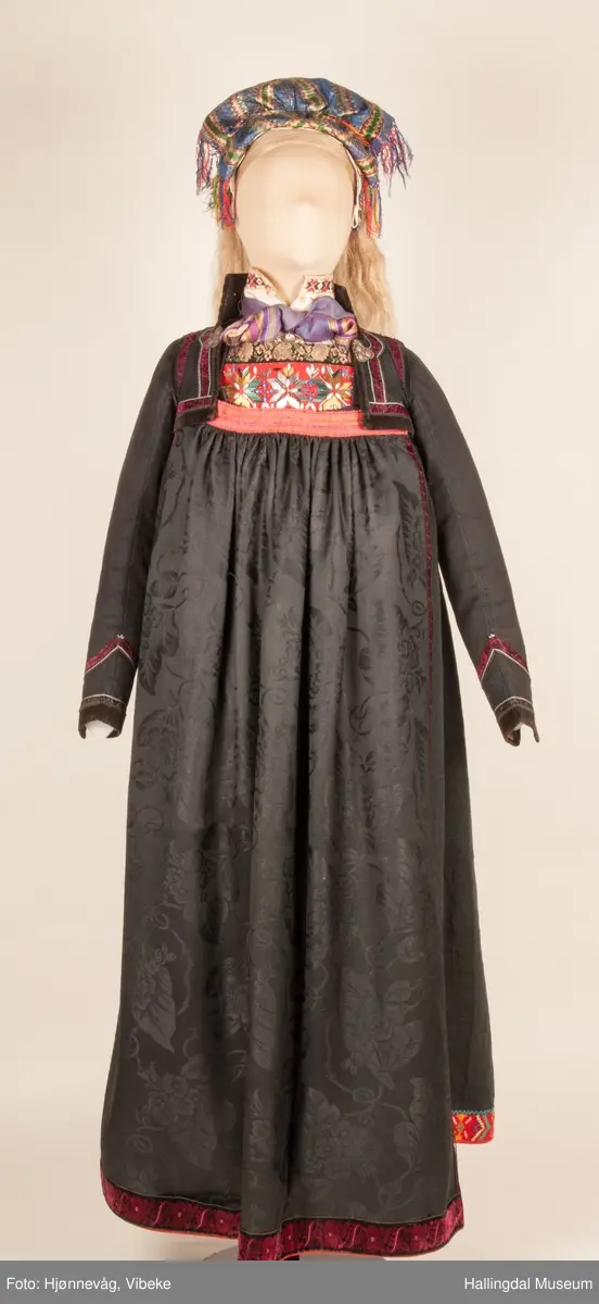 Kone fra Øvre Hallingdal i hallingbunad.  Hun bærer hette.  Kona står utstilt i den faste bunadutstillingen på Hallingdal Museum, Nesbyen.  Hallingbunaden består av mange draktdeler; trøye, Forkle HFN 02033, stakk, hette, silketørkle i halsen, perlet bringduk el. brystklut og skjorte.