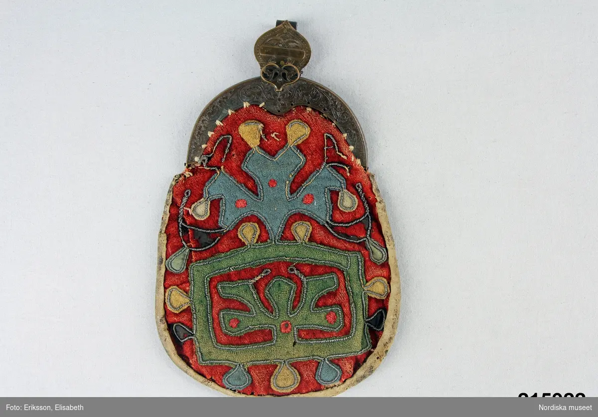 "Kjolsäck, framsida röd vadmal, applikation i olikfärgad vadmal, tennbroderier; bygel, ornerad mässing; hake mässing, märkt UDP 1814; baksida skinn."
/enligt intendent Inga Wintzells anteckningar