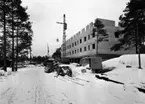 Bygge
Exteriör, byggnad under uppförande i vinterlandskap med granar. Snötäckt väg i förgrunden.