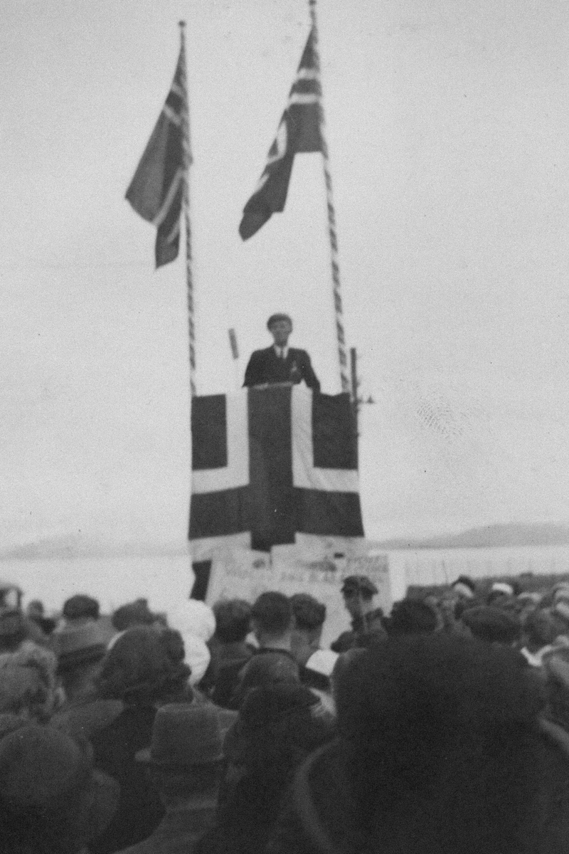 Mann på talerstol dekorert med norske flagg. I forgrunnen en folkemengde.