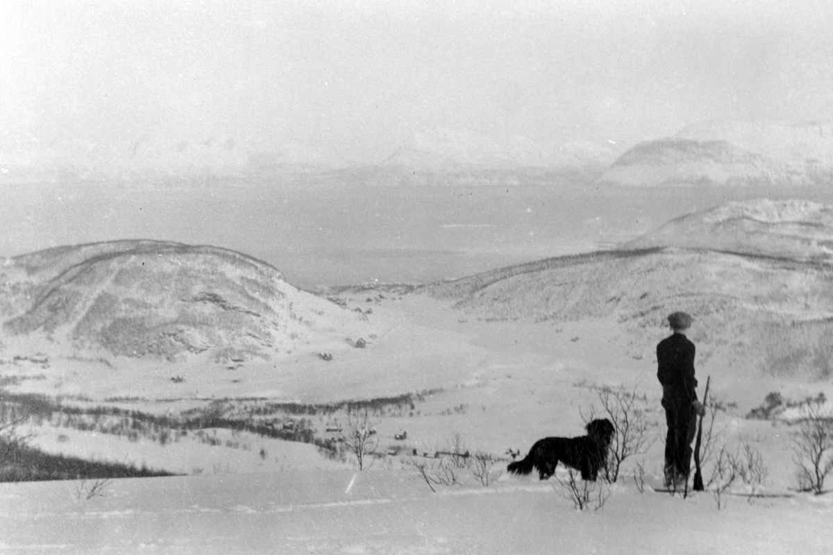 Mann med hund og gevær står i snøen på toppen av Kilhusåsen og ser utover mot Kilhus og Sama.