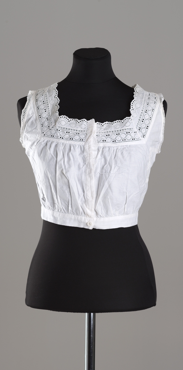 Hvitt underliv i tynt bommullslerret. Rynker over påsydd linning og på bryst. Firkantet hals dekorert med brede blonder foran og smalere blonder bak. Ermåpning kantet med smale blonder. Knappestolpe med skjult knapping, 4 knapper med hull, derav den nederste synlig. Knappene er trukket med hvitt lerret.